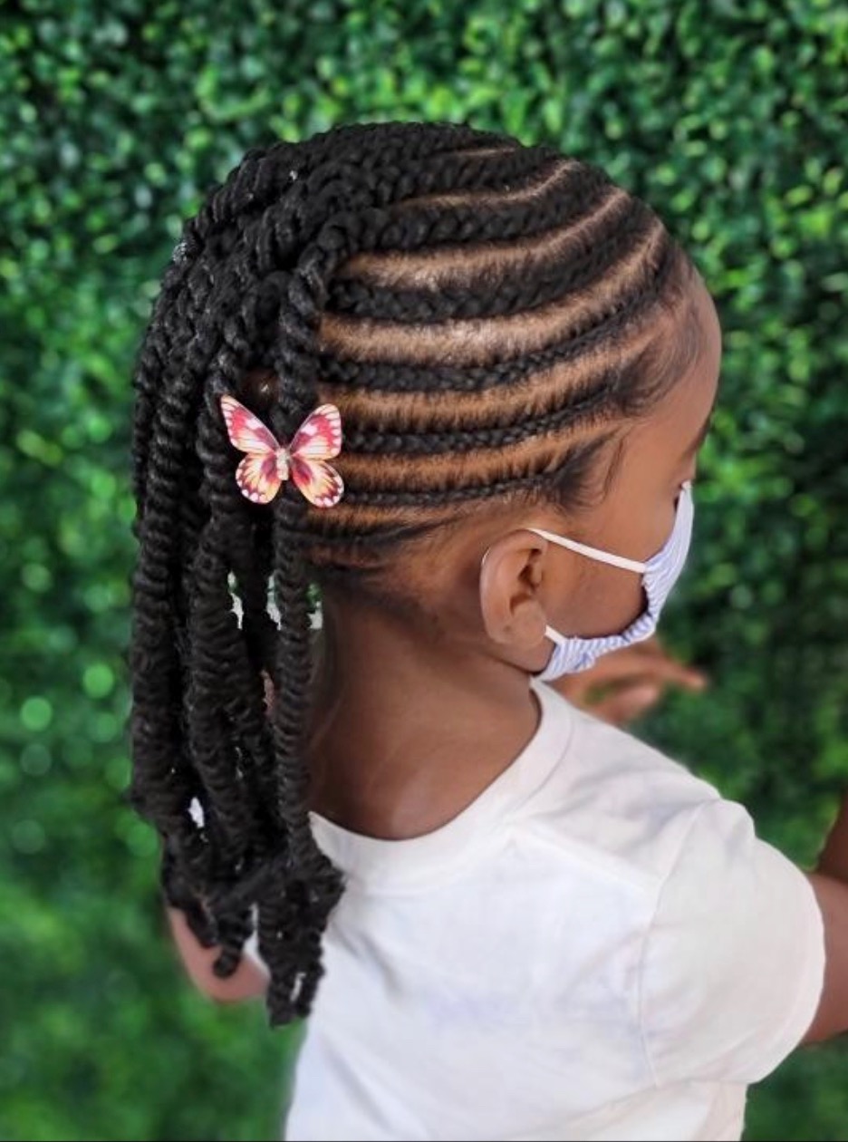 A young black girl, with cornrows that end in twisted braids. She has a pink butterfly pin in her hair.