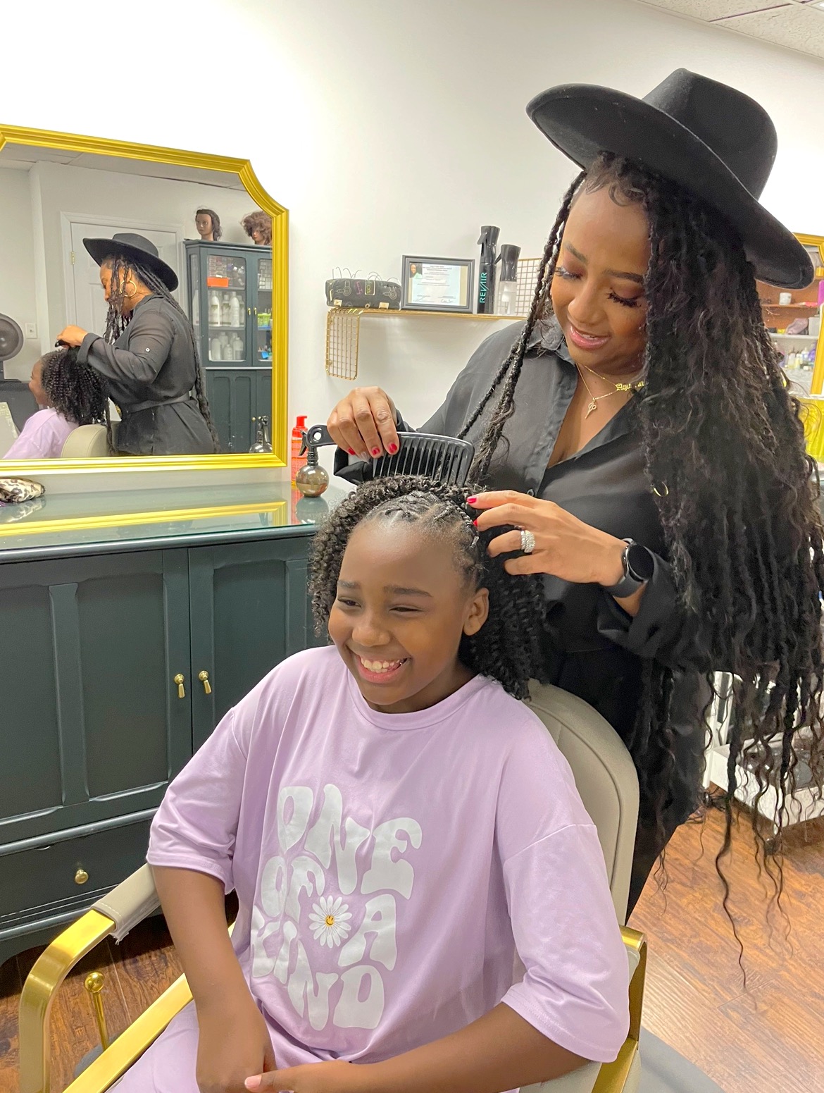A hair stylist and a young client. The stylist is black, wears a wide brimmed black hat, and has braided hair. The client is a young black girl and has half braided curled hair.