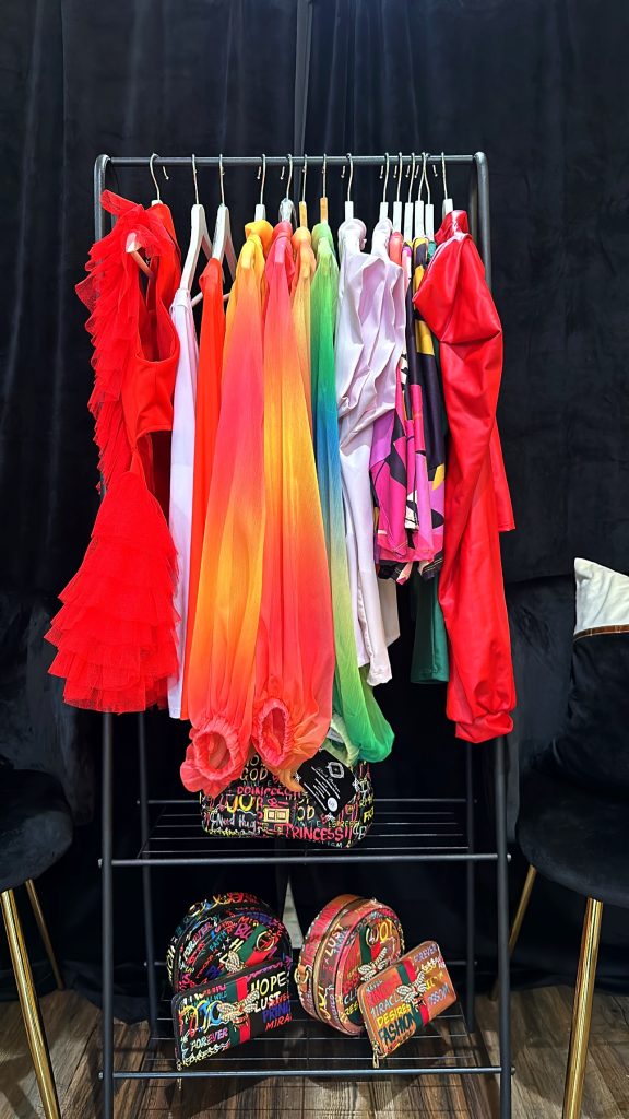 A rack and shelve, with red, white, and yellow dresses hanging. And multicolor bags on the shelf.